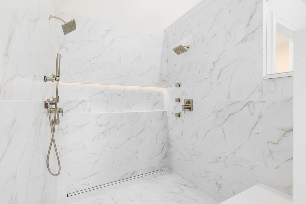 Luxurious walk-in shower with large format white marble tiled walls, gold faucets, and modern shelving with ambient lighting